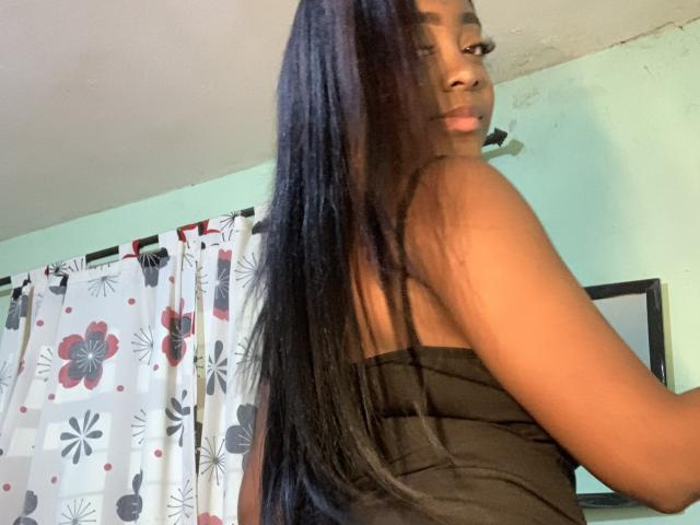 EbonnyMichell with long black hair, looking back, wearing a black strapless top, showing her brown skin and small tits, in a room with a patterned curtain.