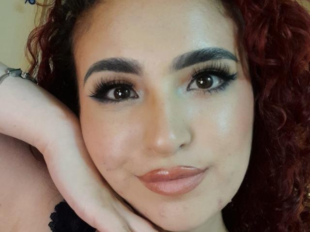 SilvanaSantos has curly red hair, thick black eyebrows, long eyelashes, and glossy pink lips. Her hand rests on her cheek, showing a soft smile. Her skin is light.