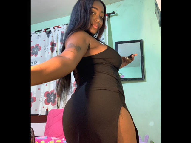 EbonnyMichell with long black hair, wearing a tight black dress with a thigh slit, showing her curvy ass and small tattoo on her arm, smiling in a green-walled room.