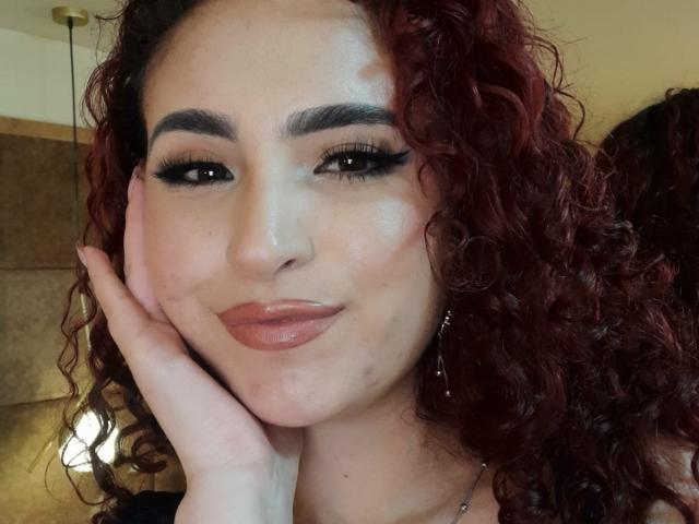 SilvanaSantos with curly red hair, dark eyebrows, and thick black eyelashes, resting her hand on her cheek, has shiny skin and pink lipstick, looking cute and confident.