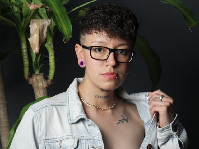 Curly-haired AllexNoa with glasses, purple ear gauges, gold necklace, and denim jacket, showing a chest tattoo and holding a shirt. Background includes green plant leaves.