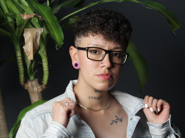 AllexNoa with curly black hair, glasses, purple ear gauges, and lip piercing, wearing a denim jacket, holding a cigarette, and tattoos on neck and chest.