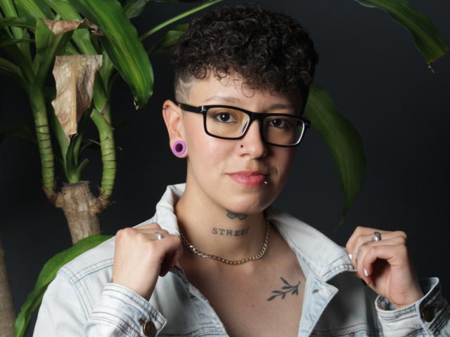 AllexNoa with short curly hair, black glasses, purple ear gauges, lip piercing, and a denim shirt, holding shirt collar, showing tattoos and necklace, standing by green plant.