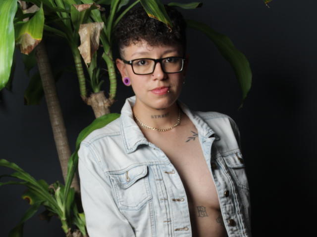 AllexNoa with curly brown hair, glasses, and purple ear gauges, wearing an open denim jacket, showing a tattooed chest, standing in front of a green plant.