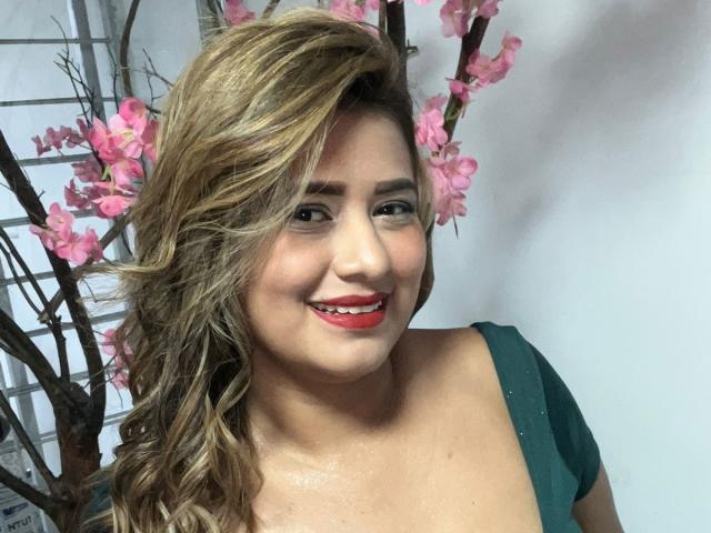SecretSexies has wavy blonde hair, red lipstick, green dress, and a cute smile. Pink flowers and tree are in the background. She's looking at the camera.