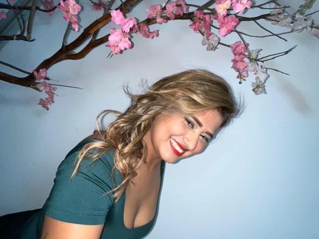 Blondie SecretSexies with wavy hair, red lipstick, and cleavage in a green dress, smiling at pink cherry blossoms. She's leaning forward, showing off her tits.