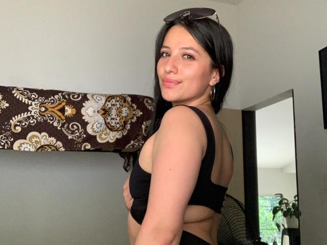 SamyNoa with dark hair, wearing black sports bra, showing sideboob, holding sunglasses on head, smiling at camera, standing in room.