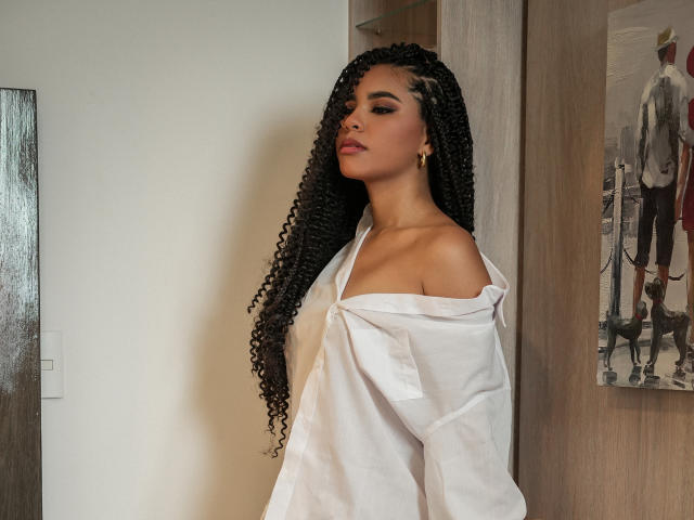 HornySweetEbony with long, curly black hair, off-shoulder white shirt, gold earrings, standing, looking slightly to the side, confident, sexy.