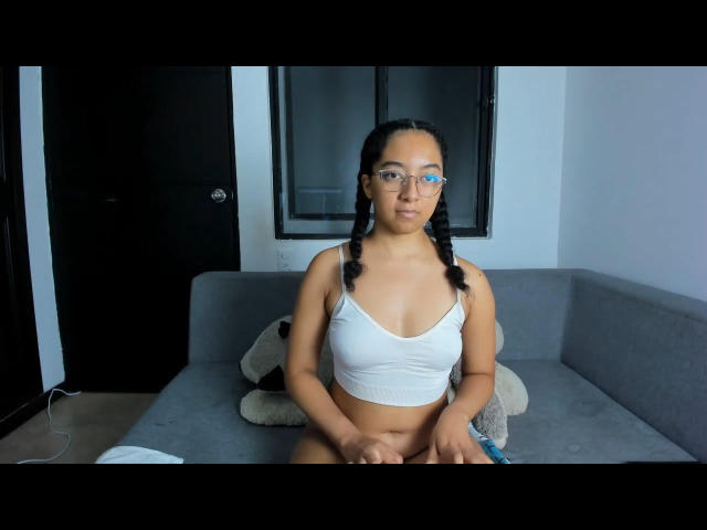 NahomiMeilleur con i capelli intrecciati, indossa un reggiseno sportivo bianco, mostrando il seno, seduta su un divano grigio, tiene in mano un telecomando blu e guarda nella fotocamera..