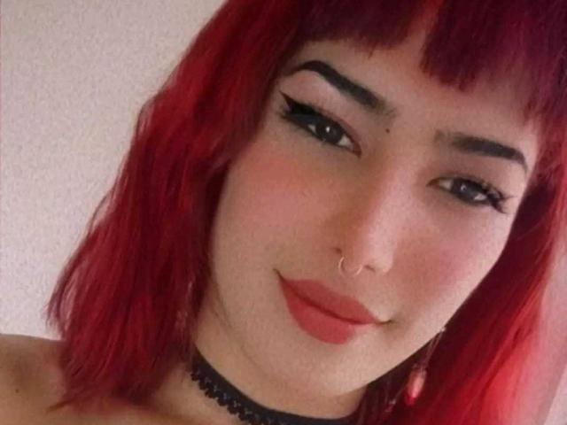 Close-up of Lunardy with vibrant red hair, straight bangs, and dark eyebrows. She has a fair skin tone, a small nose ring on her right nostril, and wears black eyeliner with a slight wing. Her lips are slightly parted with a subtle pink lipstick. She's wearing a black choker with a lace pattern. The texture of her hair is slightly wavy. Lunardy's expression is a gentle, confident smile. The image has a soft, slightly blurred quality, emphasizing the facial features and hair color. Slight red tint in the hair highlights.