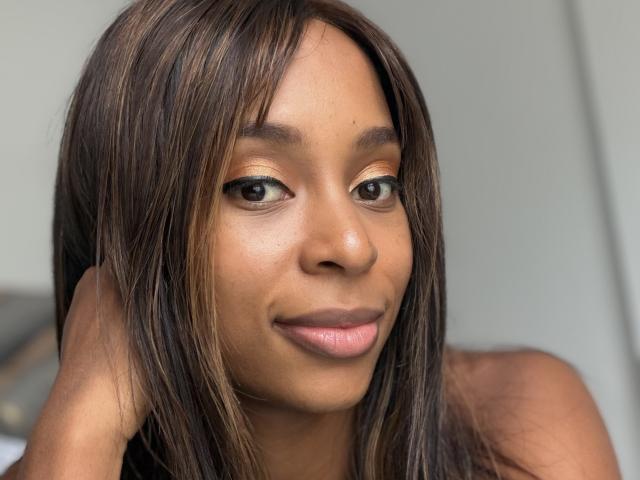 EmmaLatin has brown skin, straight brown hair with highlights, small nose, full pink lips, and brown eyes, smiling softly. She's touching her hair with her right hand.