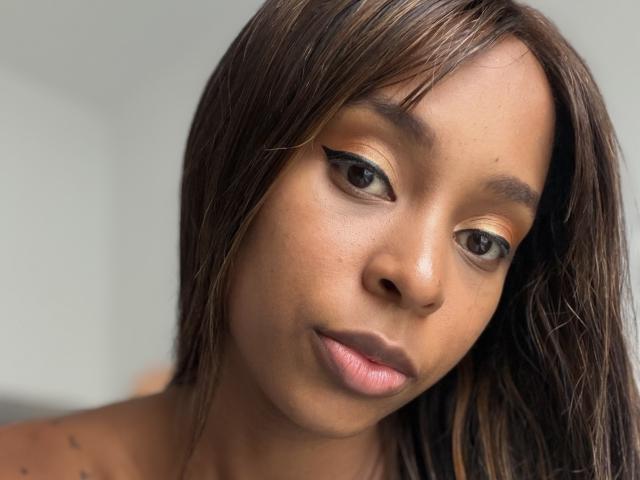 EmmaLatin's face, with brown skin, straight brown hair, and black eyeliner, looking slightly downward, has small, pink lips and a neutral expression. Her eyes are dark and slightly open.