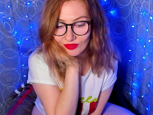PaulinaSunny has shoulder-length blonde hair and wears black-rimmed glasses. She has fair skin and red lipstick, sitting with her hand resting on her cheek. She's wearing a white t-shirt with green and red text. Blue fairy lights are in the background, creating a soft, blue glow. The curtain has a circular pattern, adding texture. The overall composition is balanced, with PaulinaSunny centered and the lights providing a contrasting color. The image is sharp with a focus on her facial expression and relaxed posture.