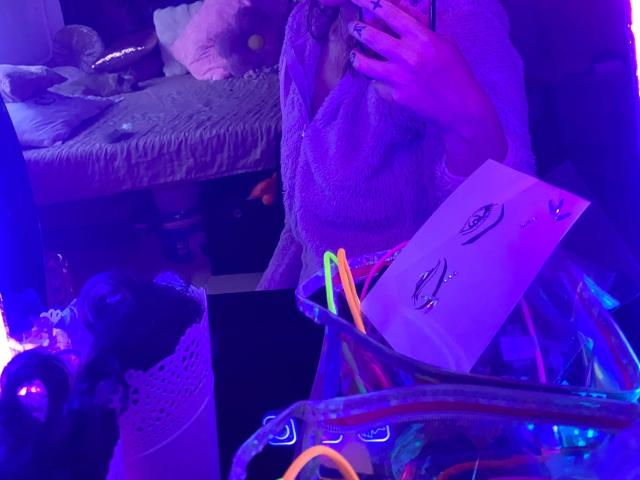 Melli-hot with purple glow, wearing a textured robe, holding a phone, mirror reflection showing colorful neon sticks and paper with heart symbols. Bed with plush toys in background.