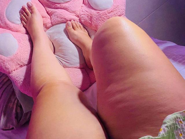 Close-up photo of MiaSp's legs on a bed. MiaSp's legs are spread apart, showing her pale skin with visible stretch marks on her inner thighs. Her toes are pointing slightly upward, resting on a pink, polka-dotted plush pillow. The pillow has a soft texture and is positioned on the left side. MiaSp's lower body is partially covered by green and yellow floral-patterned fabric. The bed has a purple sheet, and the room is lit with purple light. The image focuses on MiaSp's legs and the pillow.