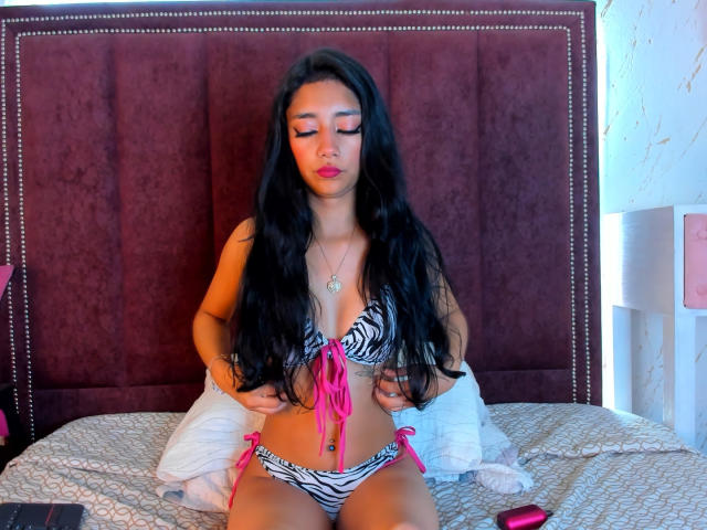 LoreneLash with long black hair, wearing a zebra-striped bikini with pink ties, sitting on a bed, hands on her belly, eyes closed, pink lipstick, white pillow behind.