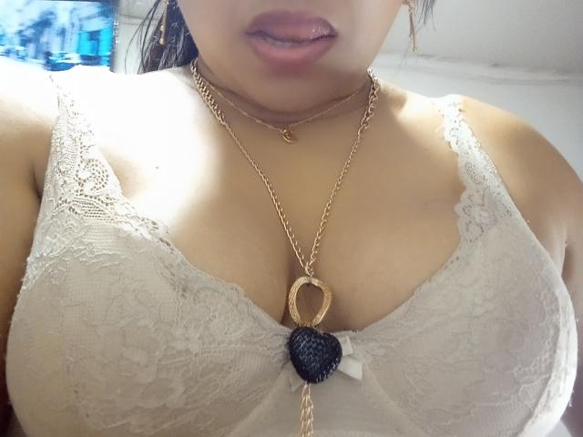 MiaSalomeChaude's tits in a white lace bra, gold necklace with heart and black berry, slightly parted pink lips. Her skin is tan, and she's wearing gold earrings.