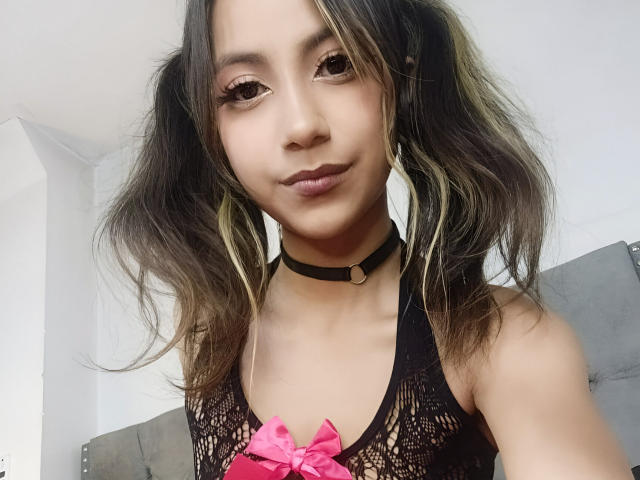 KarlaMiilan has medium brown skin, wavy dark hair with blonde highlights, wearing a black choker and lace top with pink bow, looking at camera with slight smile.