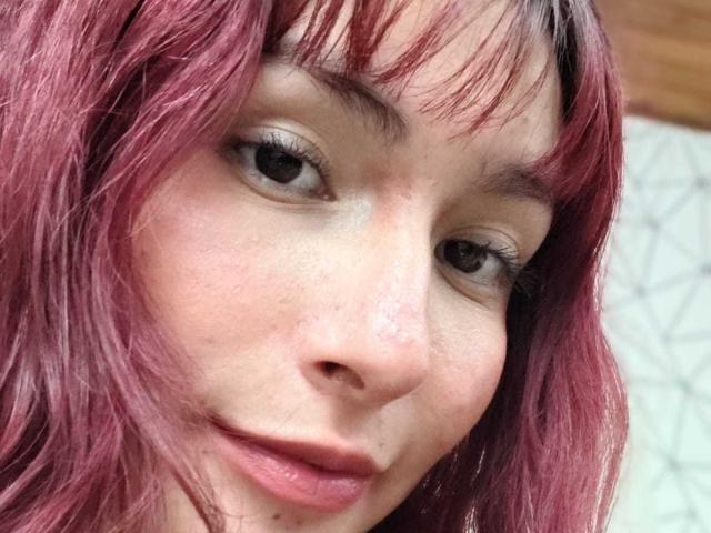 IsabellaHallad's face with pinkish-red hair, light skin, and small nose, smiling slightly with visible freckles and subtle makeup, showing her brown eyes and full lips.