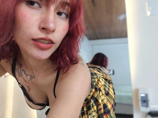 IsabellaHallad with red hair, black bra, and yellow plaid skirt, leaning forward, showing small tits, tattoo on chest, mirror reflection, slightly open mouth, in a bedroom.