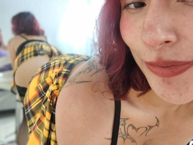 IsabellaHallad with red hair, plaid skirt, black strap, tattoos on shoulder and chest, smiling, showing her ass in mirror reflection, slightly bent over.