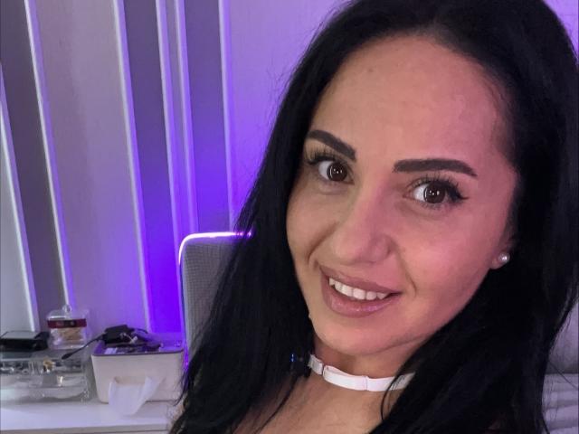 Jenniferwild69 with black hair, brown eyes, and a smile, wearing a white choker, has small tits, and is looking at the camera in a purple-lit room.