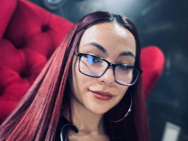 ValeryHarllow has red hair, black glasses, and a subtle smile, with hoop earrings, and a slight nose ring, sitting in front of a red, tufted chair.