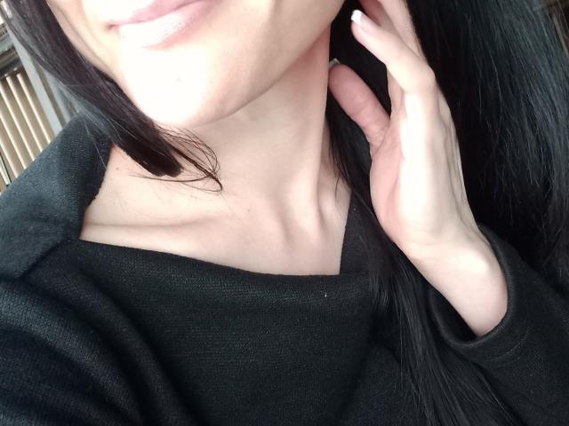 BellaJulls with pale skin, black shirt, and long black hair, touching her neck with a hand, showing a subtle smile and soft, pink lips. Her tits are small and visible.