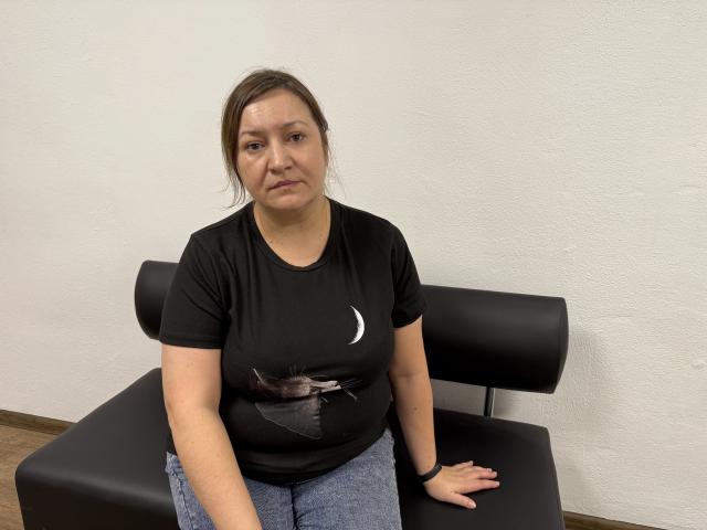 NadinForest, uma mulher corpulenta com cabelo castanho, vestindo uma camiseta preta e calças jeans azuis, está sentada em uma cadeira de couro preto, com uma expressão séria e com as nádegas levemente salientes..