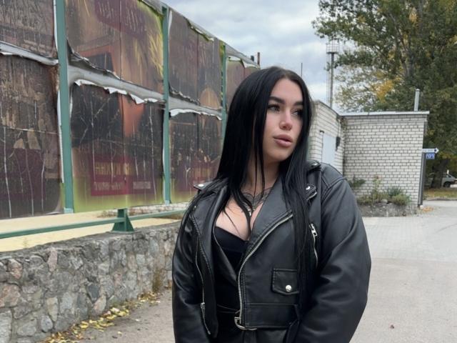 SummerMisato with long black hair, wearing a black leather jacket, black top showing her tits, standing outside, looking slightly to the side with a neutral expression.