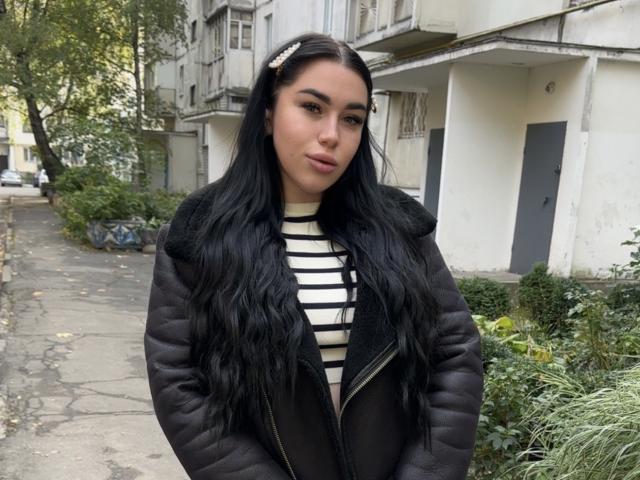 SummerMisato with long black hair, wearing a black puffy jacket and striped shirt, standing on a cracked sidewalk with green plants and old buildings in the background.