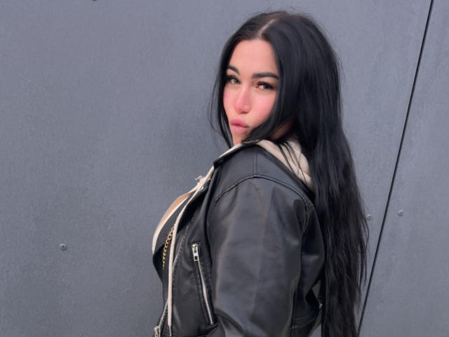 SummerMisato with long black hair, looking over her shoulder, wearing a black leather jacket, showing her ass slightly, standing against a gray wall, with a neutral expression.