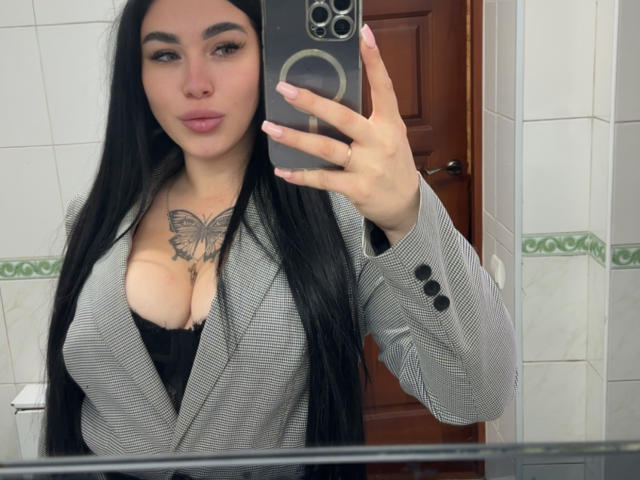 SummerMisato with long black hair, pouty lips, wearing a grey checkered blazer, black bra, showing large tits, butterfly tattoo on chest, taking a mirror selfie in a bathroom.