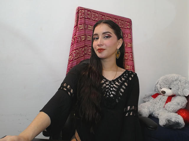 JalimaMona with long dark hair, black lace top, gold hoop earrings, sitting in chair, grey teddy bear with red ribbon on right, pink patterned pillow behind.