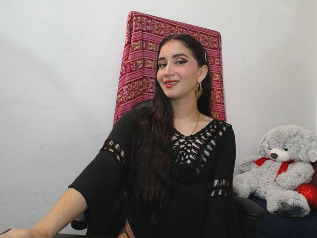 JalimaMona with long dark hair, wearing black lace top, gold earrings, smiling, sitting in chair, gray stuffed teddy with red ribbon on right, red patterned pillow behind.