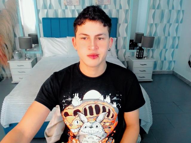 AxelSavage, com cabelo preto encaracolado curto e pequenas brincos, vestindo uma camiseta preta com um desenho animado colorido, sentado em frente a uma cama com estofamento azul. As pupilas olhando ligeiramente para o lado..