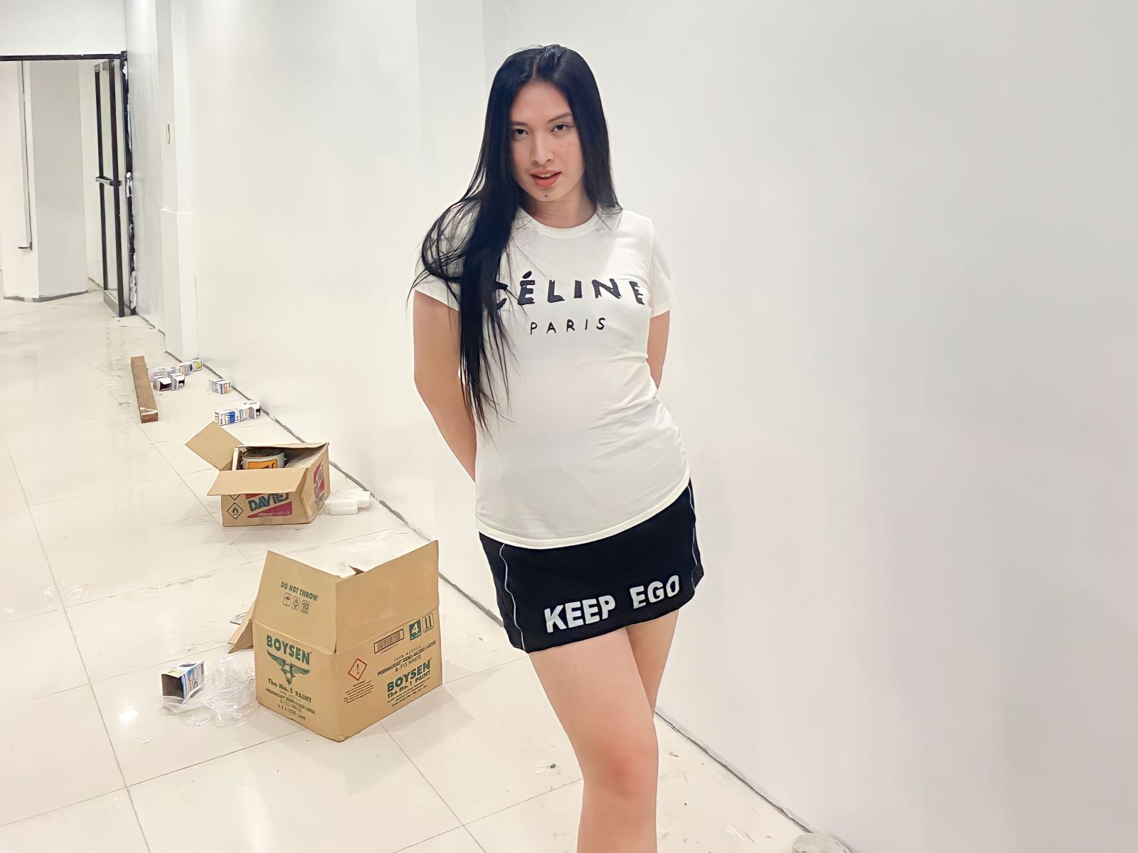 Dahlia69 stands in a bright, white hallway with her arms behind her back. She has long, straight black hair and a slight smile. She wears a white t-shirt with "CELINE PARIS" text and a black mini skirt with "KEEP GO" in white letters. Her skin is light. Several cardboard boxes are on the floor, with a few items scattered around. The image is well-lit, emphasizing the white walls and floor. Her relaxed pose contrasts with the minimalistic, almost sterile environment.