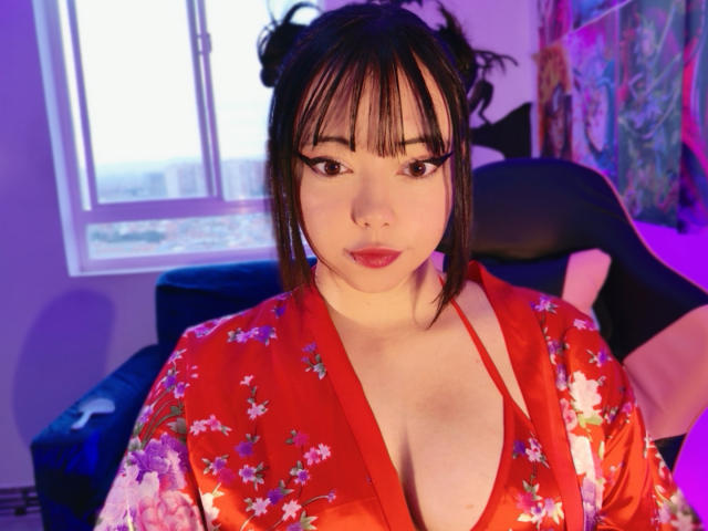 Arieyaara with Asian features, black hair in buns, red floral kimono, deep cleavage, black eyeliner, purple and blue lighting, sitting in a chair, looking at camera.