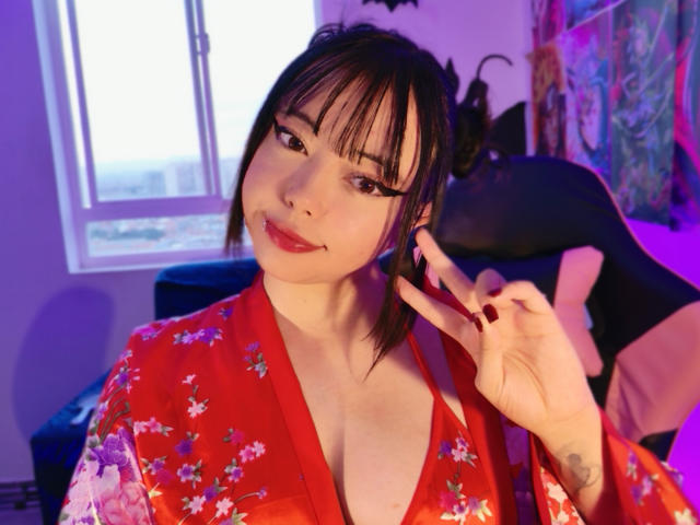 Arieyaara, con tratti asiatici, frangia nera e un kimono floreale rosso, mostra il suo seno con un sorriso appena accennato, facendo un segno di pace, sotto luci viola e blu..