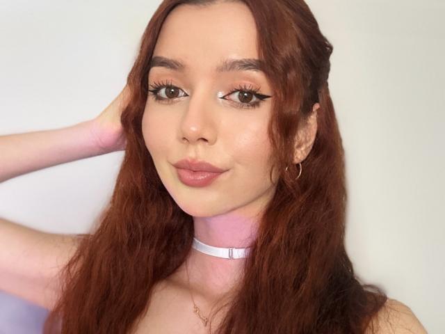 DreamyLovee with wavy red hair, brown eyes, and pink lips, wearing a white choker and gold hoop earrings, smiling softly. Her hand rests behind her head.