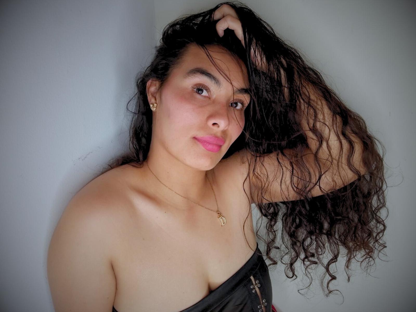 DahianRoss with long, curly black hair, pink lipstick, and tan skin, wearing a strapless black top, hand in hair, necklace with small pendant, smiling slightly.