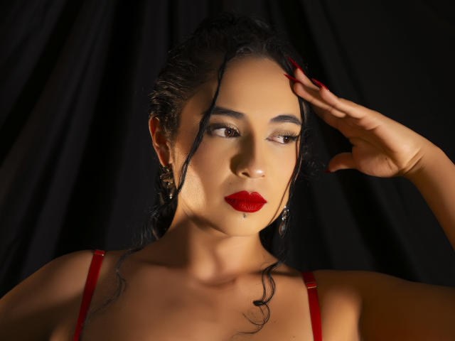 LilithStygian heeft donker haar, rode lippenstift en rode nagels, draagt een rode band over haar schouder, met haar hand tegen haar hoofd, op een zwarte achtergrond. Ze kijkt naar de zijkant..