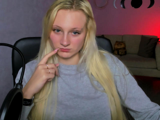 MdamForulis with long blonde hair, pink nail, grey sweater, and finger on lips, sitting in black chair, looking at camera, red light in background, soft pink skin.