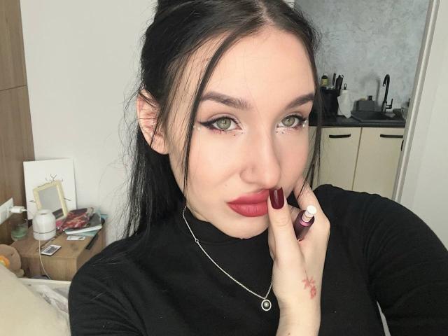 NikkiGoldX with green eyes, red lipstick, black dress, and red nail polish, touching her lip with a finger, wearing a necklace, in a bedroom, showing a slight smile.