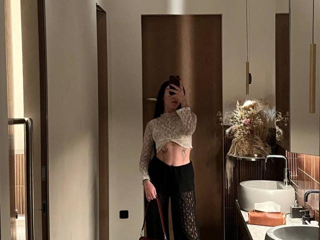 NikkiGoldX with long black hair, holding phone, wearing lace top revealing abs and tattoo, black fishnet pants, standing in bathroom, mirror selfie, showing small tits, slim, sexy.