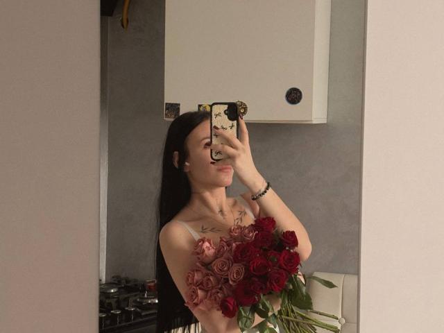 NikkiGoldX with long black hair, holding a bouquet of red and pink roses, taking a mirror selfie, wearing a strapless top, smiling, and showing off her tits.