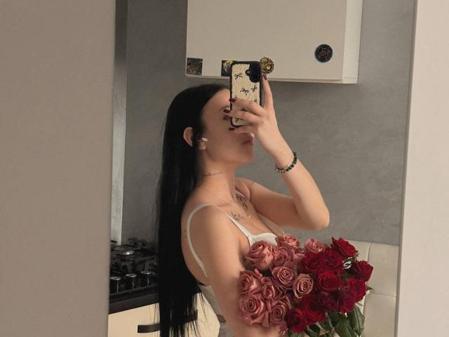 NikkiGoldX with long black hair, holding roses and red roses, wearing a white bra, taking a mirror selfie in a kitchen, phone with floral case, earrings, bracelet.