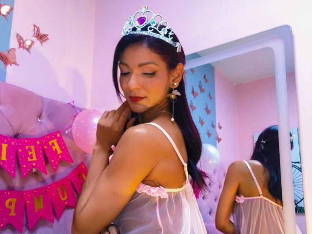 HannaPretty with dark hair, red lipstick, and pink lingerie, wearing a sparkly tiara, looking back with a slight smile, standing in front of a mirror. Boobs visible through sheer fabric.