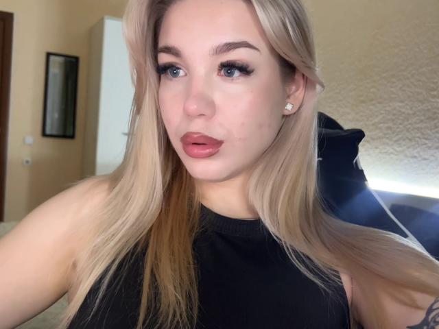 WildMaksi has blonde hair, blue eyes, and wears a black sleeveless top with pink lipstick. Her nose is slightly pinkish. She's looking at the camera with a neutral expression.
