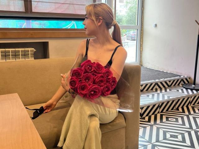 WildMaksi with pink roses, holding bouquet in transparent bag, sitting on beige chair, wearing black bra, white pants, blonde ponytail, holding black shoes, on geometric patterned floor.